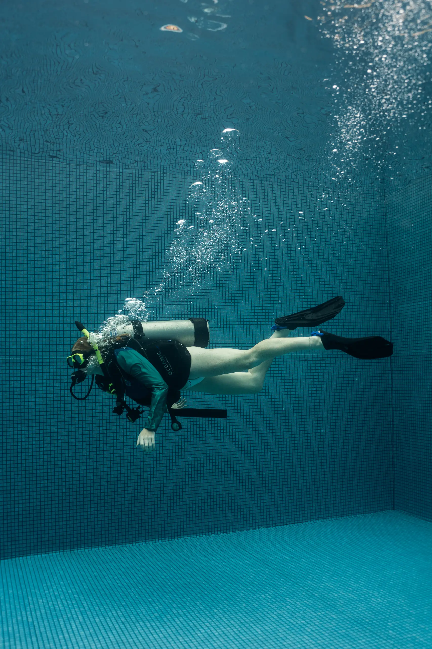 A diver gliding along the bottom of the training pool, tank on the back and bubbles rising
