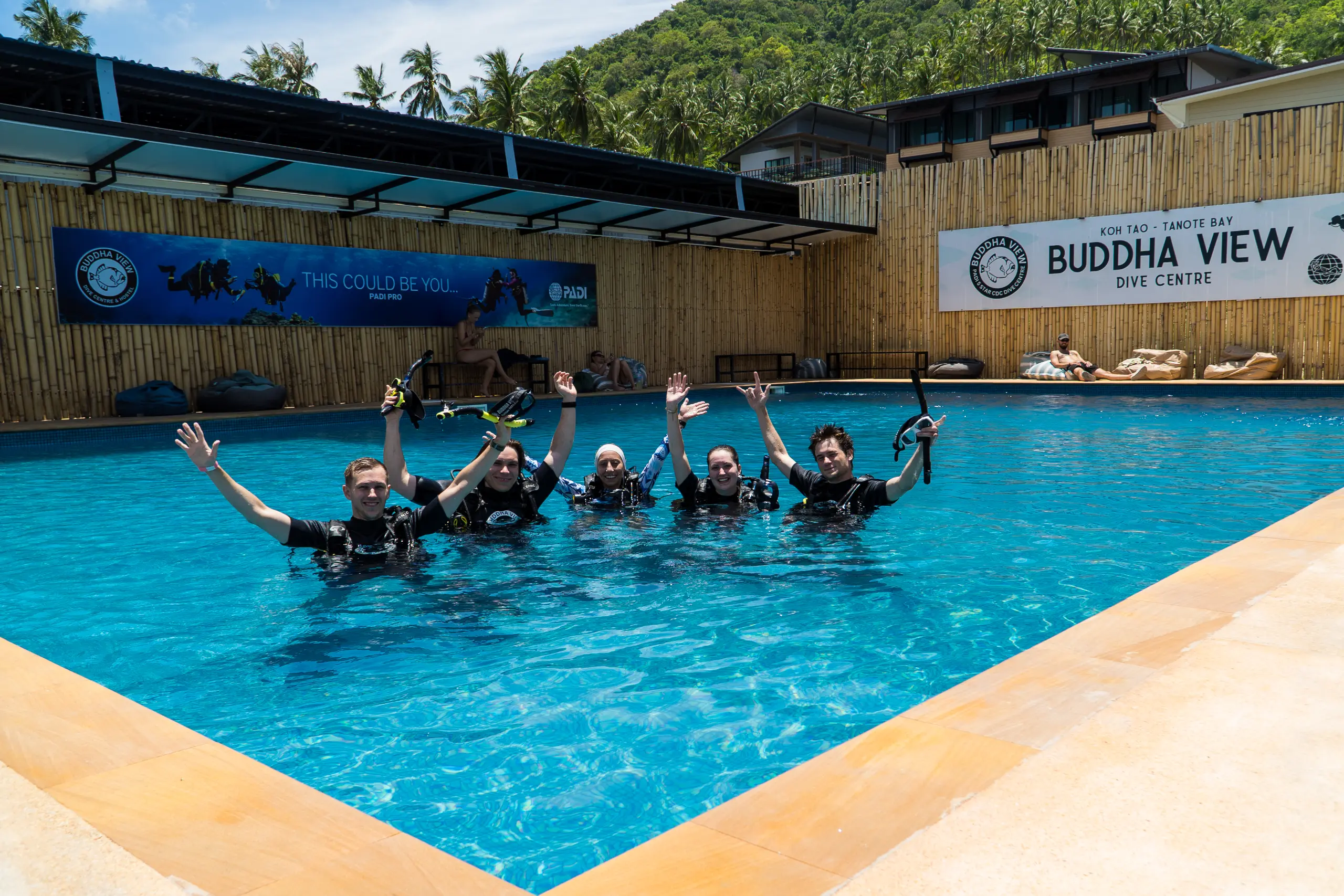 Five students in wetsuits standing in the Buddha View training pool, arms raised with their fins and snorkels, the dive centre sign behind them